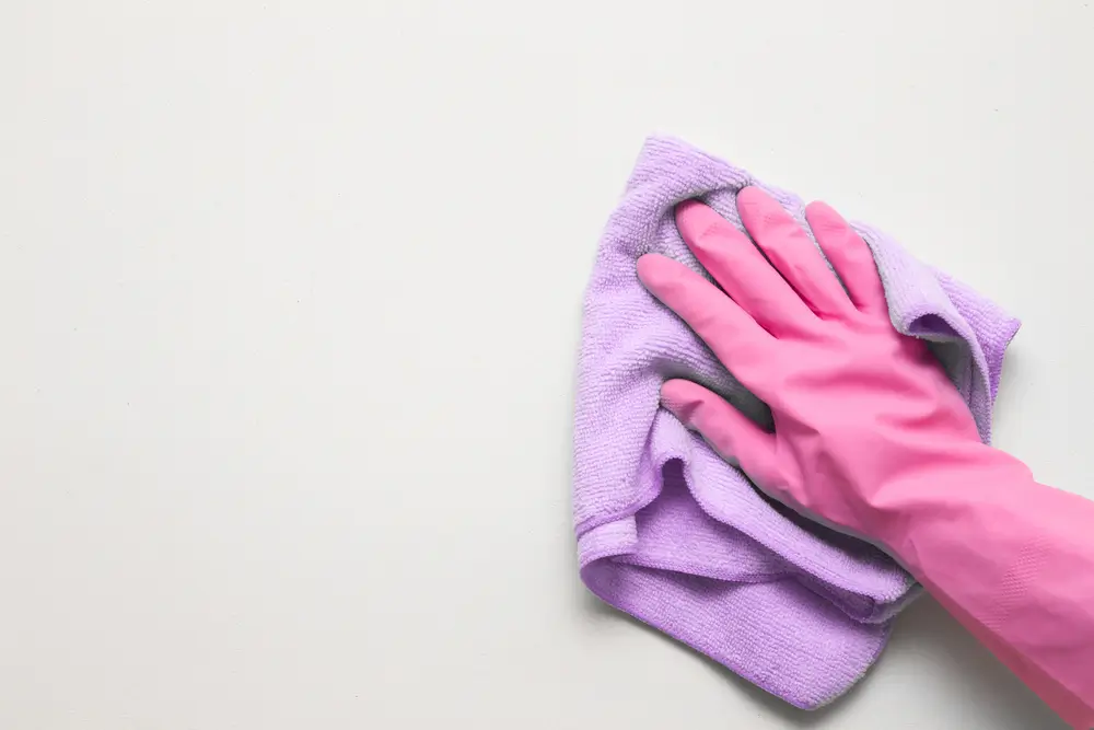 Ręka damska w różowej rękawicy ochronnej wycierająca biała ścianę z kurzu suchą szmatką.