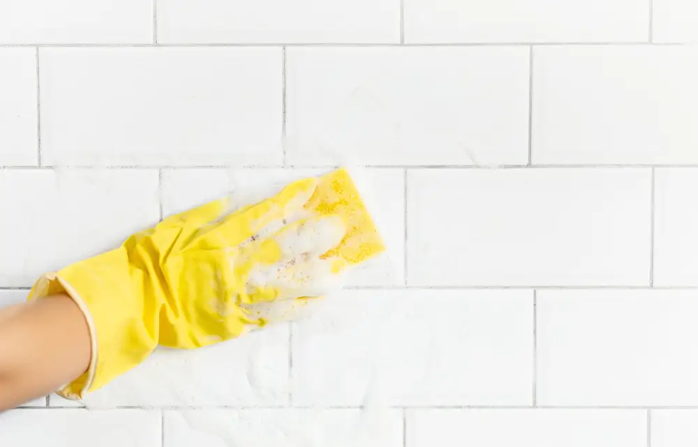 Damska ręka w żółtej rękawiczce, czyszcząca białe fugi przy pomocy gąbki