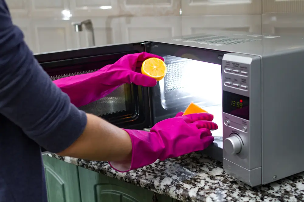 Kobieta w różowych rękawiczkach na dłoniach, czyszcząca mikrofalówkę przy pomocy gąbki i cytryny. Sposób na to, jak wyczyścić mikrofalówkę.